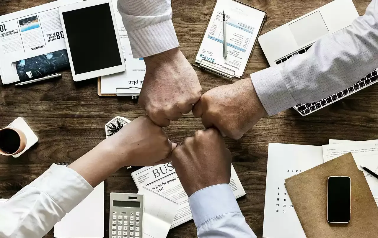 Über einem Tisch mit Arbeitutensilien treffen sich fünf Mitarbeiterfäuste.